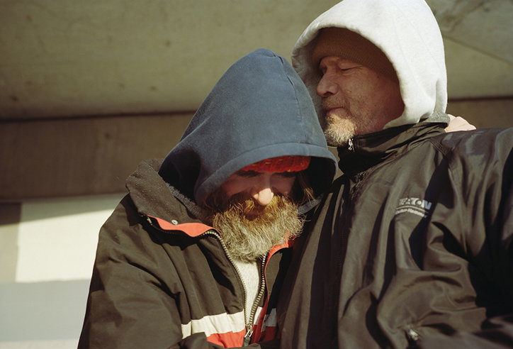 Two people lean into each other, wearing heavy winter coats and hoods. One person has a full beard and a red beanie tucked under a navy hood.