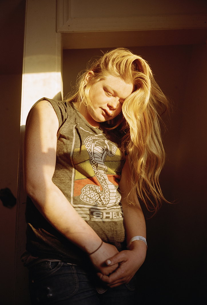 A person with long blonde hair stands in a doorway, partially lit by a beam of sunlight. They wear a grey T-shirt featuring a graphic of a cobra and the word “SHELL”.