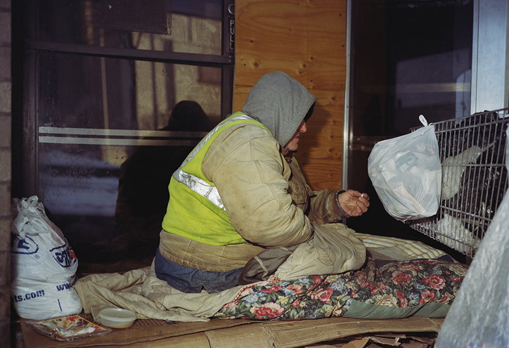 A person wearing a grey hoodie and a high-visibility yellow vest sits on a makeshift bed of cardboard and floral blankets. They face a small wire cage containing a bird.