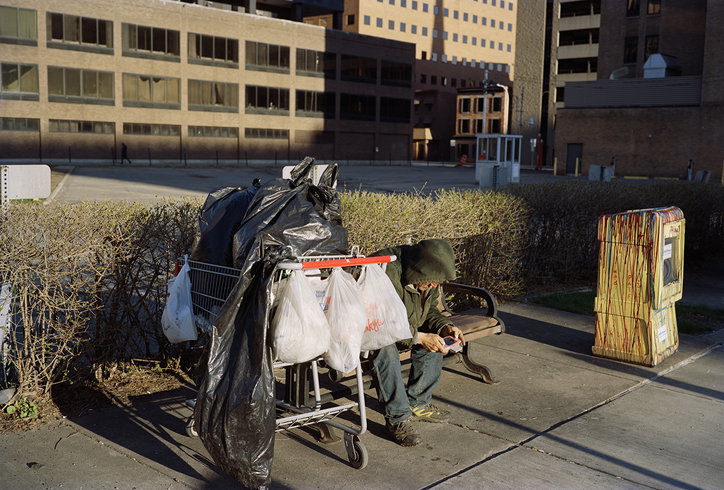 A person sits on a park bench next to a shopping trolley piled with black plastic bags and white plastic sacks. They are looking down at a handheld device in their lap.