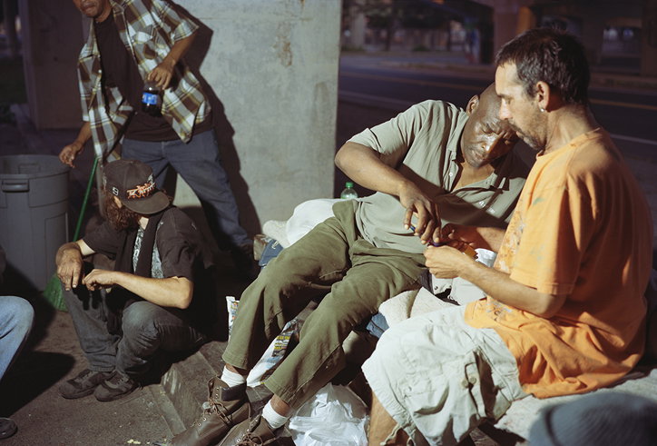 Three people gather on a city pavement at night. One person leans back against a wall while another sits beside them, focused on something in their hands. A third person stands nearby, wearing a checked shirt.