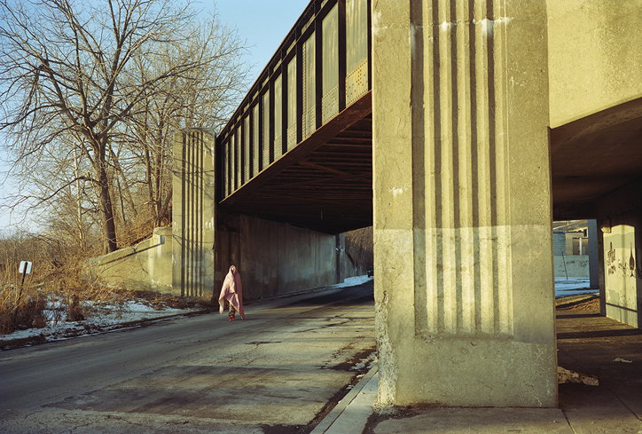 A person draped in a pale pink cloth walks along a road beneath a concrete bridge. To the left, bare winter trees stand against a clear blue sky.