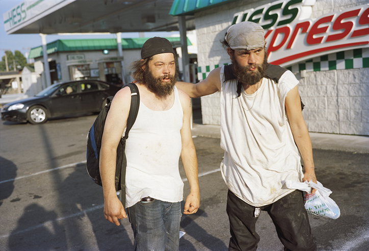 Two people with long beards stand outside a Hess Express petrol station. They wear white vests and head coverings, with one person resting an arm on the other's shoulder.