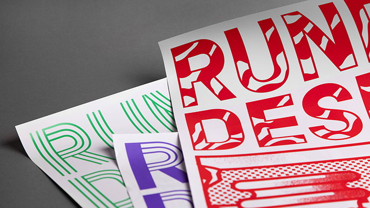 Three printed posters overlap on a grey surface. The top poster features bold red lettering that reads “RUN” and “DES”, with a patterned fill inside the characters. Other posters in green and purple show similar typographic styles.