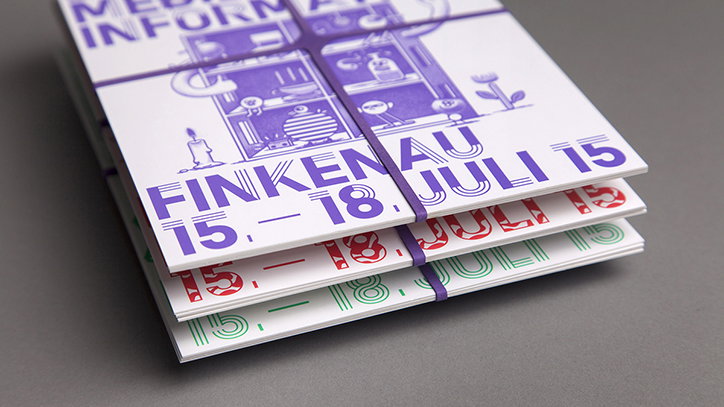 A stack of printed booklets tied with purple elastic bands. The top cover features purple illustrations and text that reads “FINKENAU 15. – 18. JULI 15”.