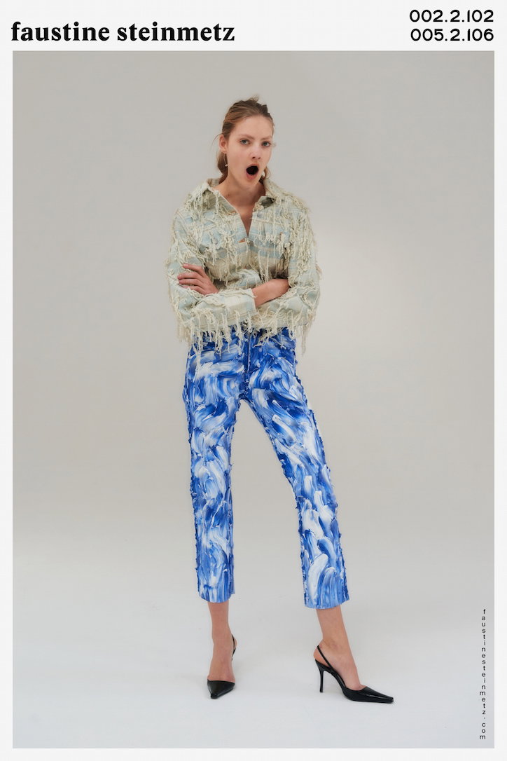 A person stands against a plain white background with a surprised expression, mouth open. They wear a fringed, pale green jacket and blue and white marbled trousers with black pointed heels. Above the image, text reads “faustine steinmetz” alongside the numbers “002.2.102” and “005.2.106”.