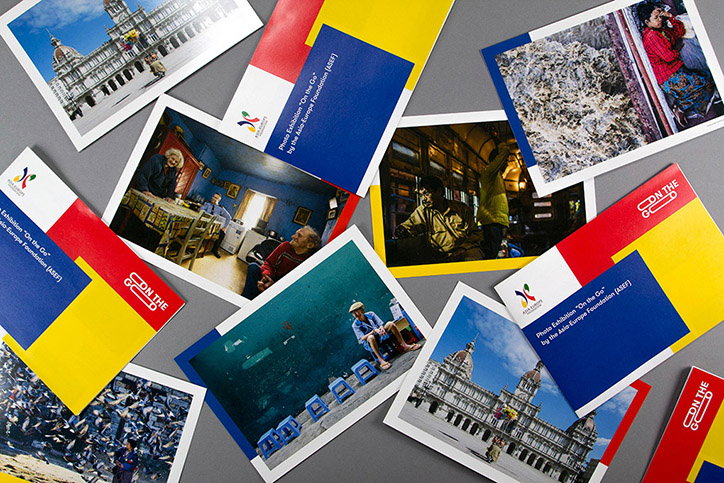 A collection of postcards scattered across a grey surface. Some show photographs of people and architecture, while others feature a bold red, yellow and blue design with the text “Photo Exhibition ‘On the Go’ by the Asia-Europe Foundation (ASEF)”.