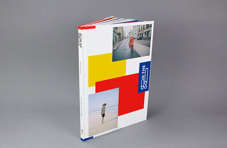A white hardcover book stands angled against a grey background. The cover features primary coloured blocks and two photographs, with the title “On the Go” printed vertically.