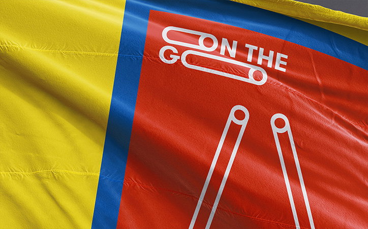 A close-up of a fabric flag with yellow, blue and red sections. A white graphic on the red section features two safety pins and the words “ON THE GO”.