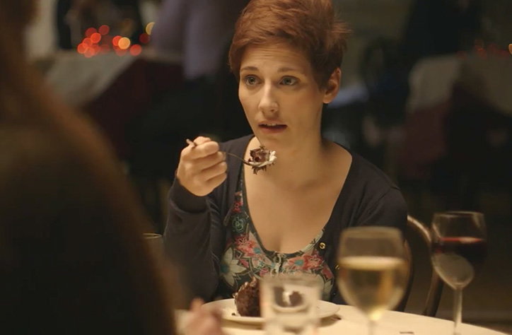 A person with short red hair sits at a restaurant table, holding a spoonful of chocolate cake. They look towards someone off-camera with a questioning expression.