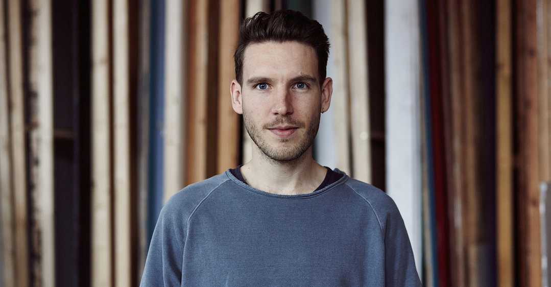 A person in a blue sweatshirt looks directly at the camera. Behind them, several vertical wooden planks stand side by side in a blurred row.