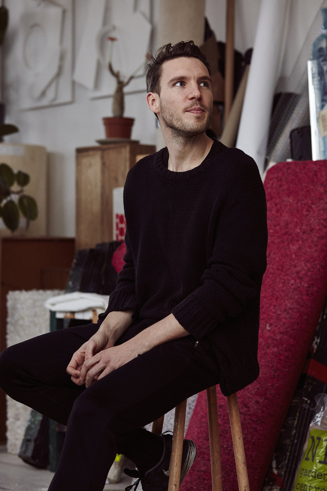 A person wearing a dark knit sweater and trousers sits on a wooden stool in a cluttered studio. They look off to the side, surrounded by art supplies, canvases and a potted plant.
