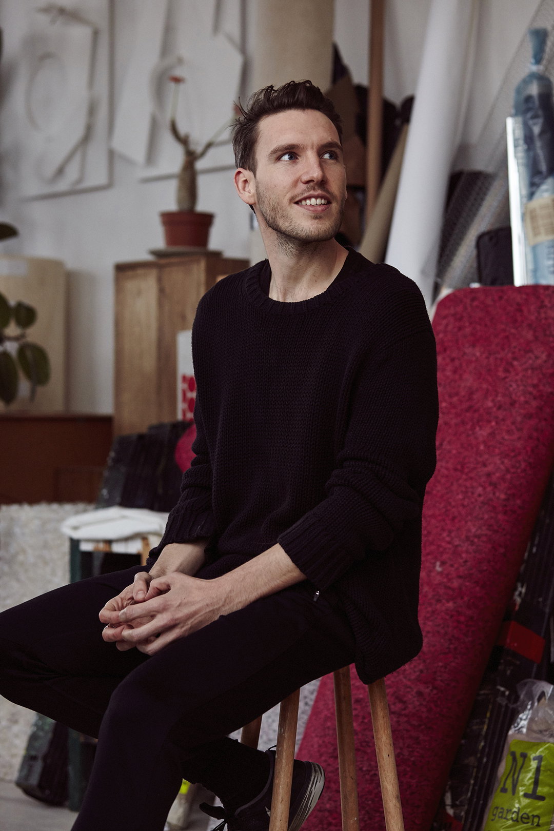 A person sits on a wooden stool in an art studio, looking off-camera with a slight smile. They wear a dark, oversized knit jumper and matching trousers, surrounded by sculptures and art materials.