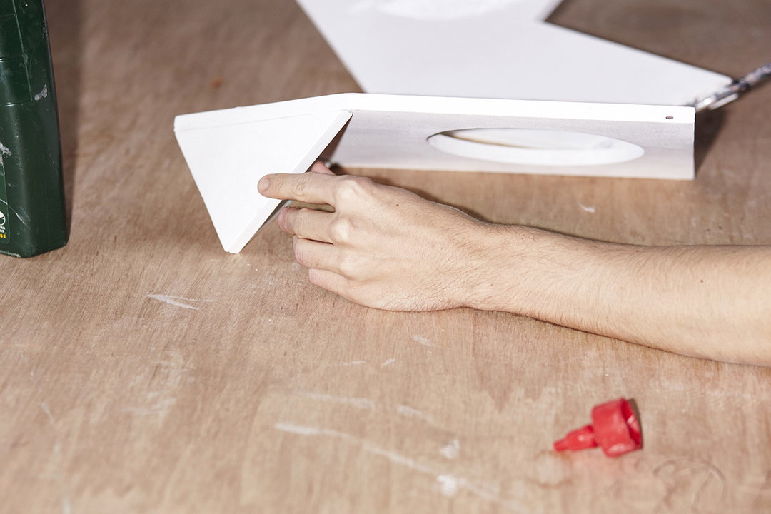 A person’s hand steadies a white, angular model on a wooden workbench. The model features a sharp fold and a circular cutout.