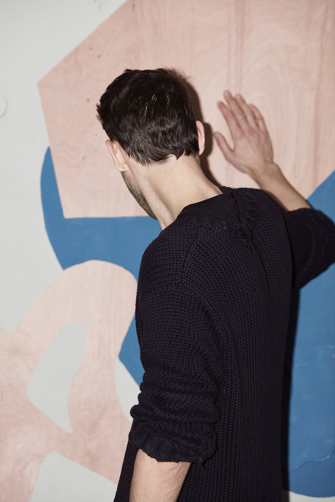 A person seen from behind stands with one hand pressed against a wall. The wall is covered in large, abstract shapes of pale pink and blue.