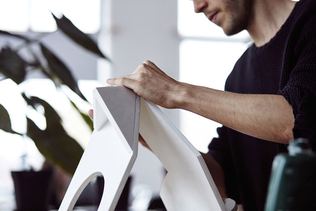 A person in a black sweater rests their hand on a white, geometric wooden structure. The object has sharp angles and a circular cutout, set against a bright, blurred background with a houseplant.