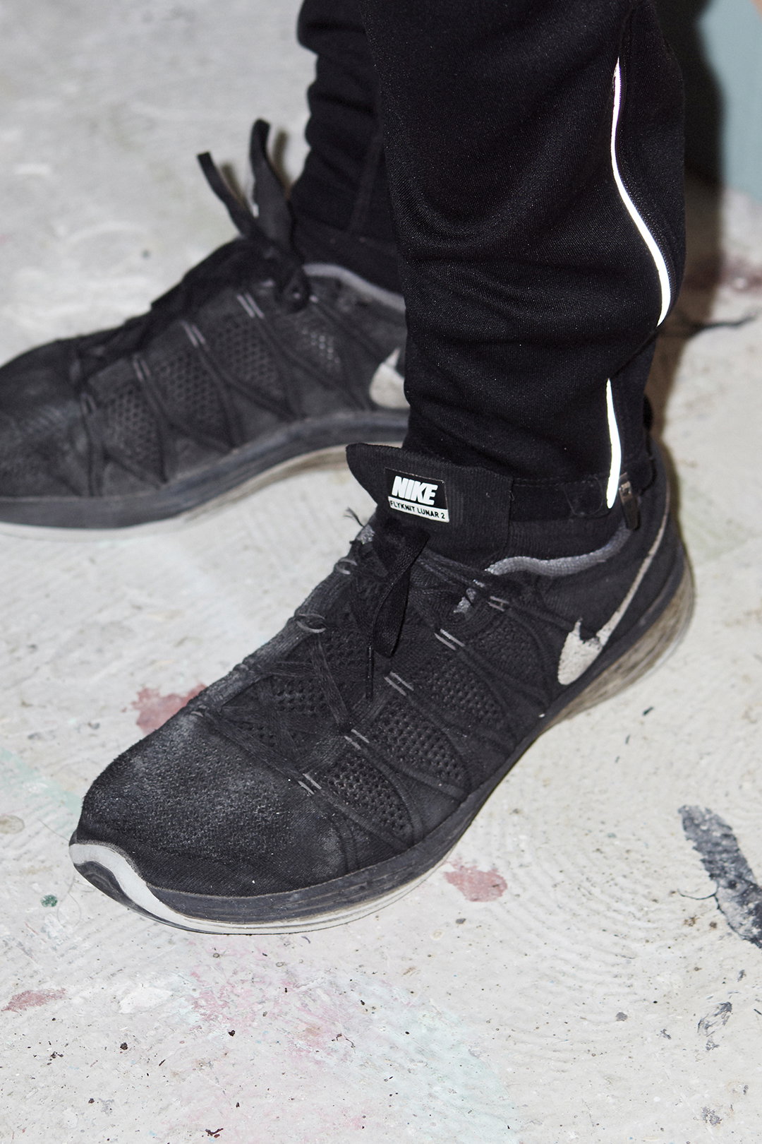 A person wears black Nike trainers and black trousers. The shoes are worn and dusty, with the “Nike” logo visible on the tongue.