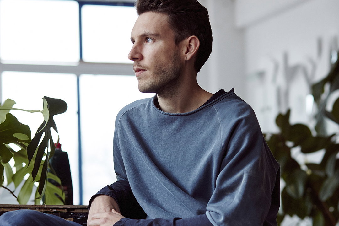 A person in a blue sweatshirt looks off-camera in a bright, airy room. Large windows and leafy houseplants frame the scene.