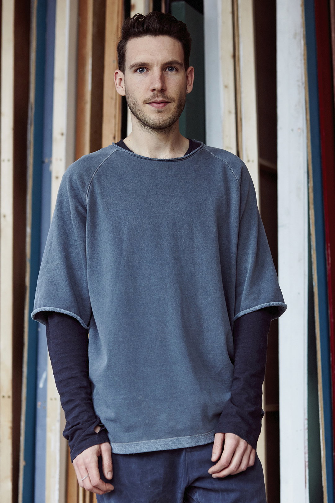 A person stands facing the camera, wearing an oversized, slate-blue top layered over a dark navy long-sleeved shirt. They are positioned against a backdrop of vertical wooden beams in various colours.
