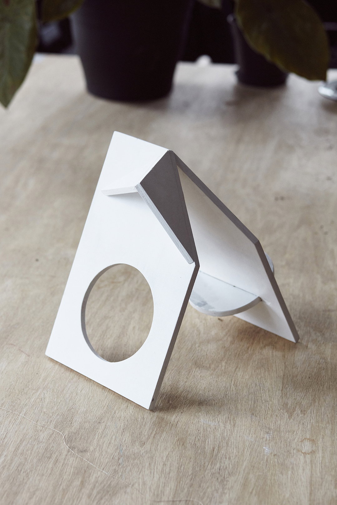 A small, white architectural model of a house with a circular cutout in the front wall. It sits on a light wood surface, with a blurred black plant pot in the background.