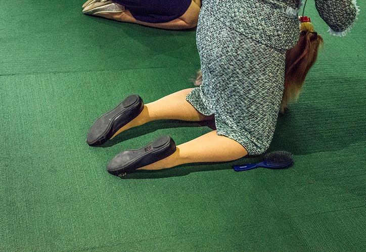 A person in a textured grey dress kneels on a green carpet. A blue hairbrush lies on the floor beside them.