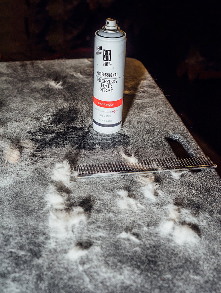 A can of “Professional Freezing Hair Spray” stands on a dark, textured surface. A metal comb and clumps of white hair are scattered around the base of the can.