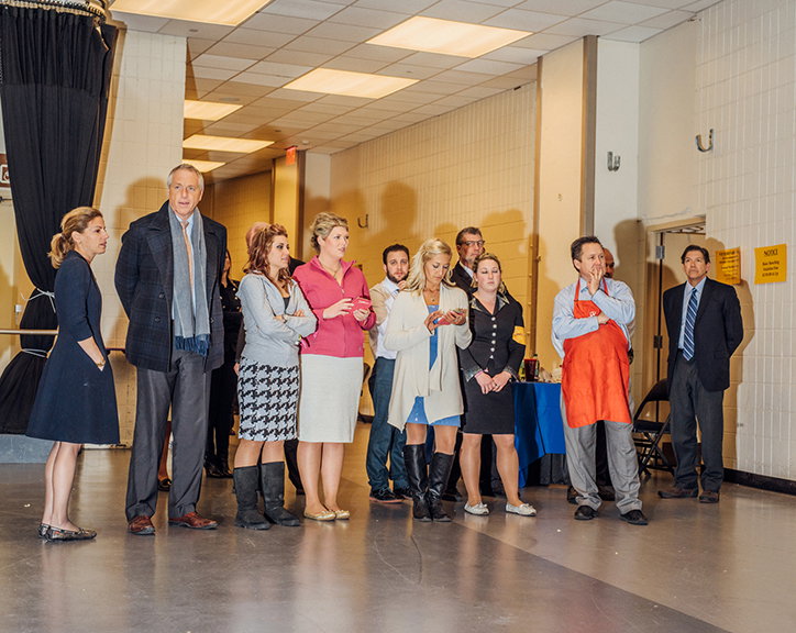 A group of people stand in a line in a brightly lit hall, looking towards something off-camera. They are dressed in a mix of smart-casual and formal attire, including one person wearing a bright orange apron.