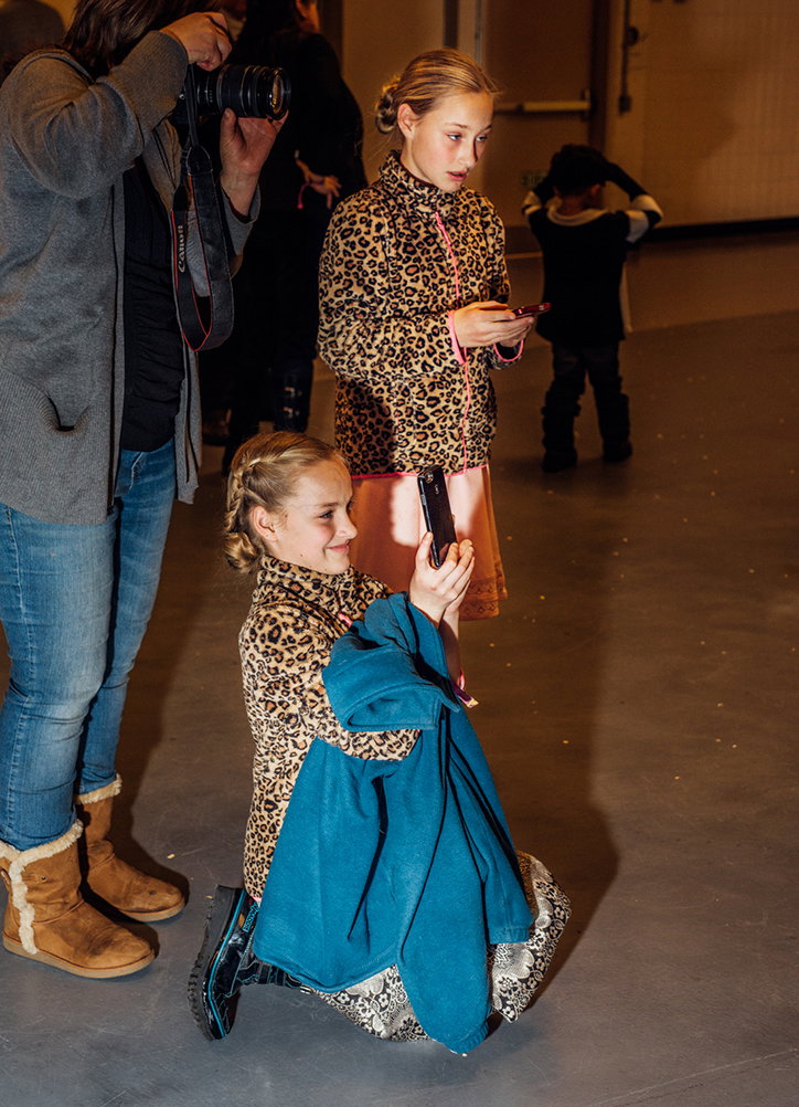 Two people wear matching leopard-print tops in a studio. One person kneels on the floor taking a selfie, while the other stands behind them looking at a phone.