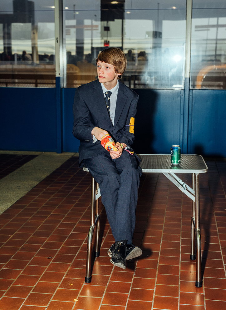 A person in a dark navy suit and tie sits on the edge of a metal table. They hold a snack bag in their hands and look off to the side.