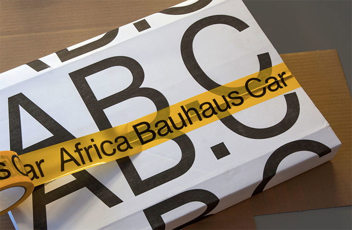 A white box is wrapped in yellow adhesive tape that reads “Africa Bauhaus Car”. Large, black, sans-serif letters “ABC” are printed across the surface of the packaging.