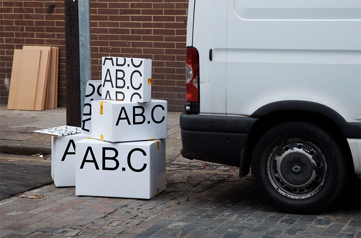 A stack of white cardboard boxes sits on a cobbled street next to a white van. Each box is printed with the bold black text “AB.C”.