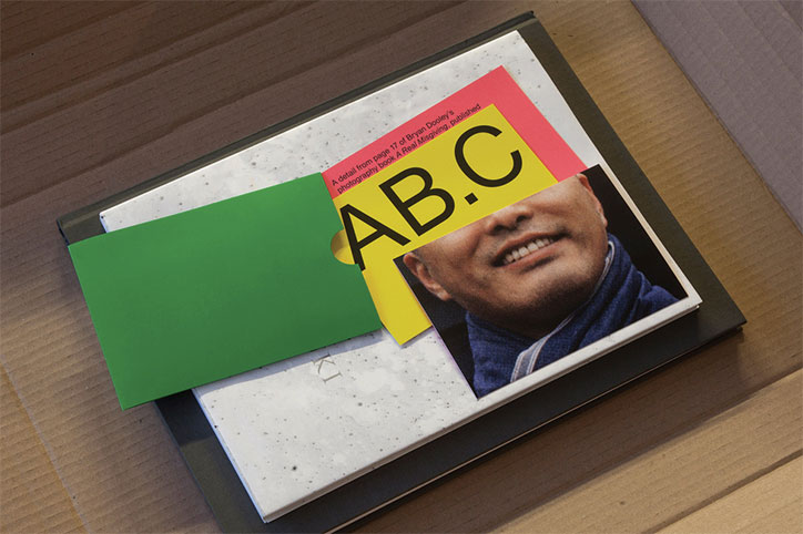 A book with a speckled white cover lies on a cardboard surface. A collage is laid over the front, featuring a person’s smiling face, a green card, and bold yellow text that reads “AB.C”.