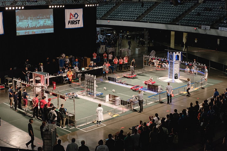 A wide shot of a robotics competition in a large arena. Teams of people stand around a fenced perimeter, watching robots compete on a grey playing field. A screen and a banner reading “First” hang above the scene, while a crowd watches from the stands.