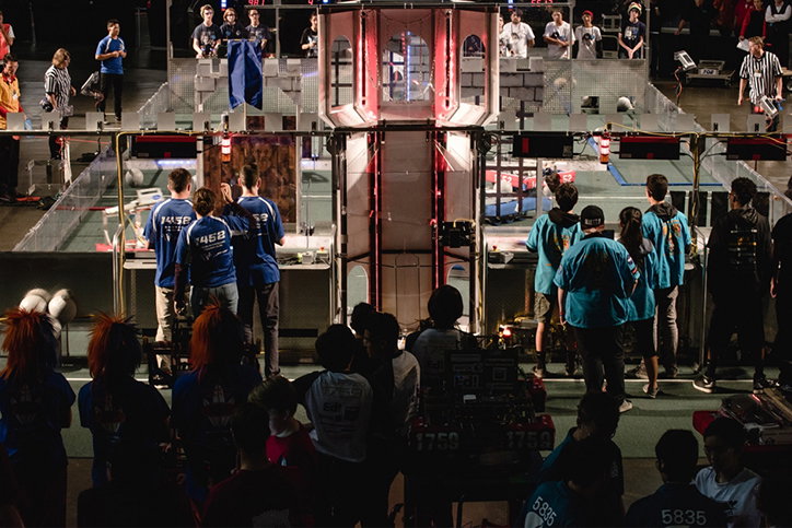 A group of people in blue team shirts stand around a robotics competition arena. In the centre, a tall, metallic structure rises above the playing field as teams prepare their robots.