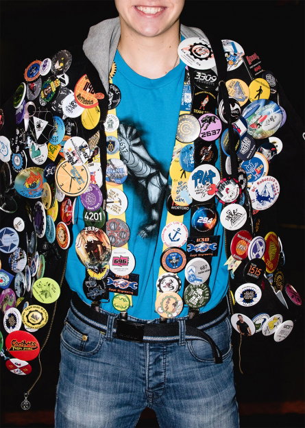 A person wears a black jacket completely covered in hundreds of colourful circular badges. Underneath, they wear a bright blue T-shirt with a graphic print and blue jeans.