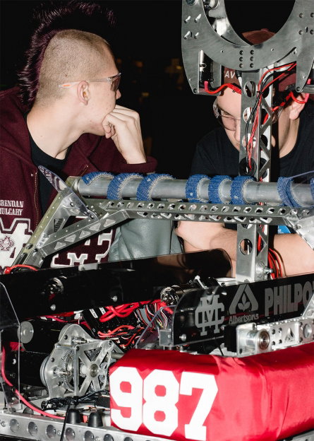 Two people lean over a complex robot made of metal frames, wires and gears. A red fabric bumper on the front of the machine displays the number “987” in white.