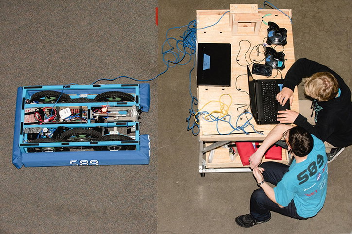 A blue robotic vehicle sits on a grey floor, connected by blue cables to a wooden workbench. Two people crouch by the bench, operating laptops and game controllers to manage the machine.