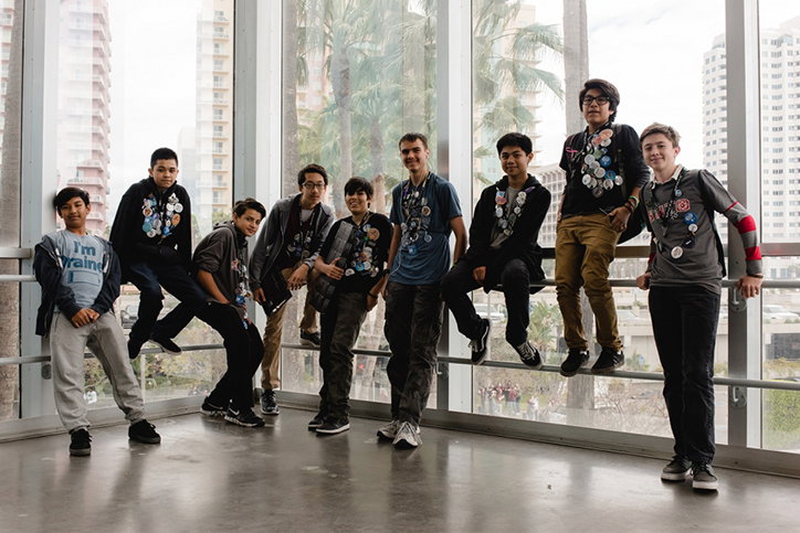 Nine people pose together in a bright, glass-walled room with city buildings visible outside. Many of them wear dark clothing adorned with numerous circular badges.
