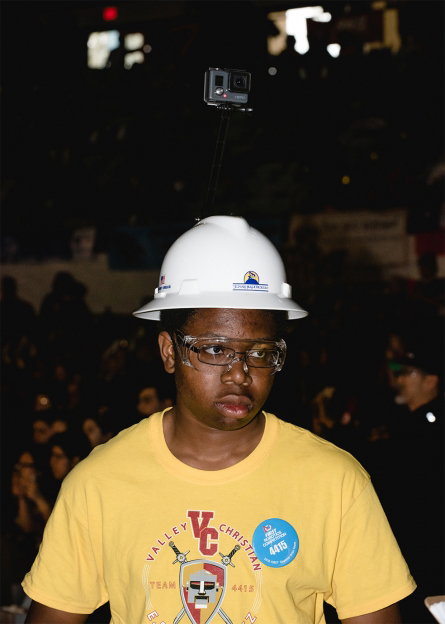 A person wearing a white hard hat and safety glasses looks thoughtfully to the side. A small camera is mounted on a tall pole attached to the top of their hat. They wear a yellow T-shirt with a logo that reads “Valley Christian”.