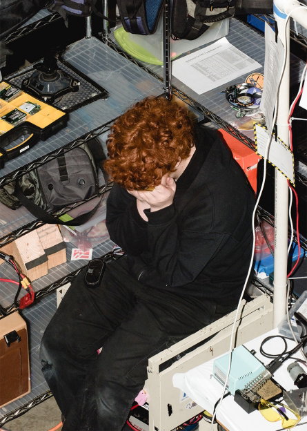 A person with curly red hair sits in a cluttered workspace, head resting in their hand. They are surrounded by electronic equipment, cables and bags on metal shelving.