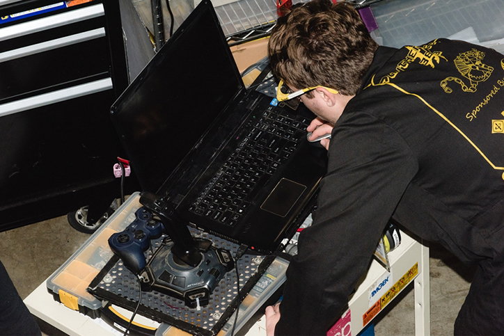 A person in black overalls leans over a laptop, looking closely at the screen. The laptop is mounted on a technical rig alongside a game controller.