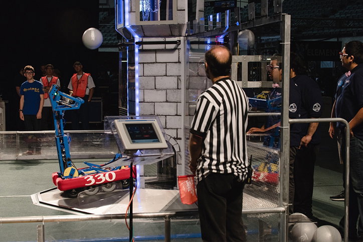 A person in a black and white striped referee shirt watches a blue and red robot on a competition floor. The robot is positioned in front of a white brick-patterned tower, with the number “330” visible on its side.