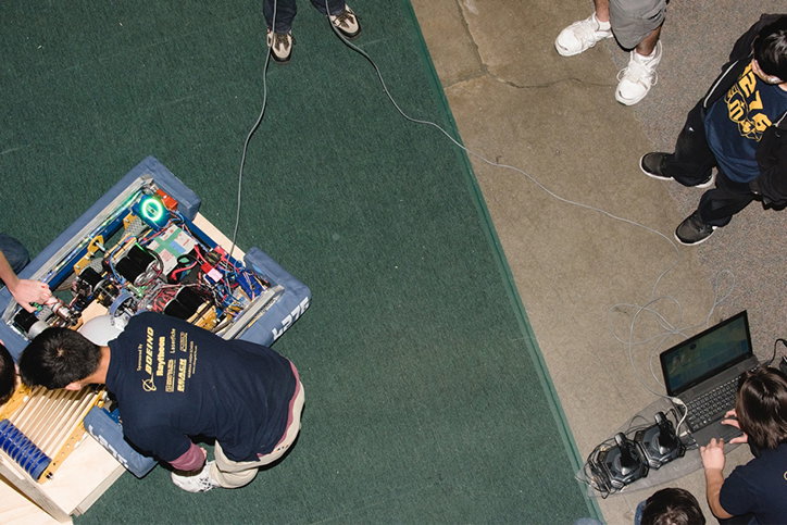 A person leans over a complex piece of electronic machinery housed in a blue plastic case. Nearby, another person works at a laptop connected to the device by a long grey cable.