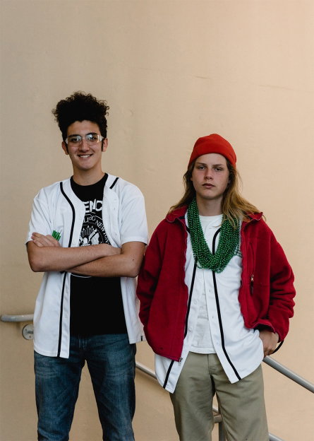 Two people stand side by side against a plain beige wall. One wears a white short-sleeved shirt over a black T-shirt and blue jeans, while the other wears a red fleece jacket, a red beanie and green beaded necklaces.