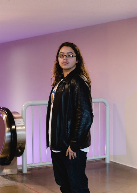 A person with long hair and glasses stands in a laundrette, wearing a black leather jacket and dark trousers. They look directly at the camera against a pale pink wall.