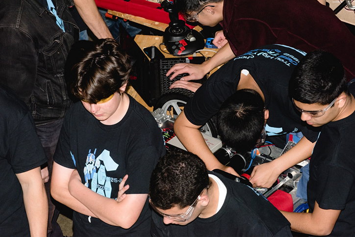 Four people huddle around a small robotic vehicle, focusing on its internal components. They wear black T-shirts and glasses as they work together to adjust the machine.