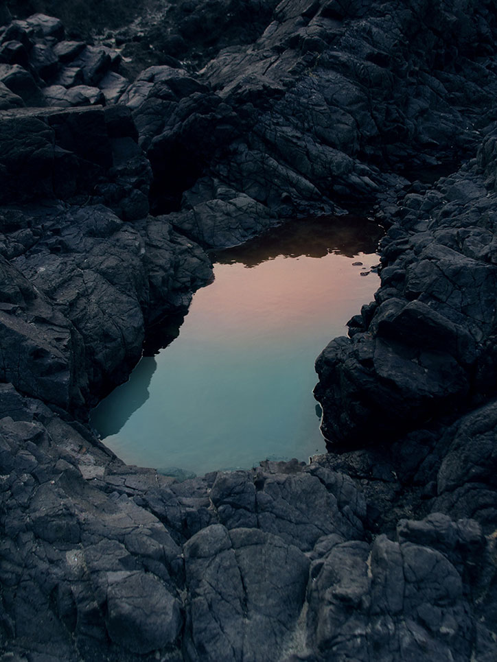 A small, still pool of turquoise water is nestled within a landscape of dark, jagged volcanic rock. The water's surface reflects a soft pink and orange glow from the sky.