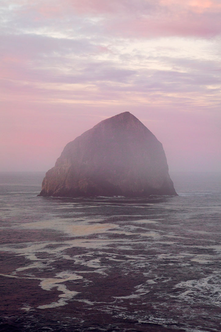 A large, rounded rock formation rises from a dark, choppy sea. The scene is bathed in a soft pink and purple haze under a cloudy twilight sky.