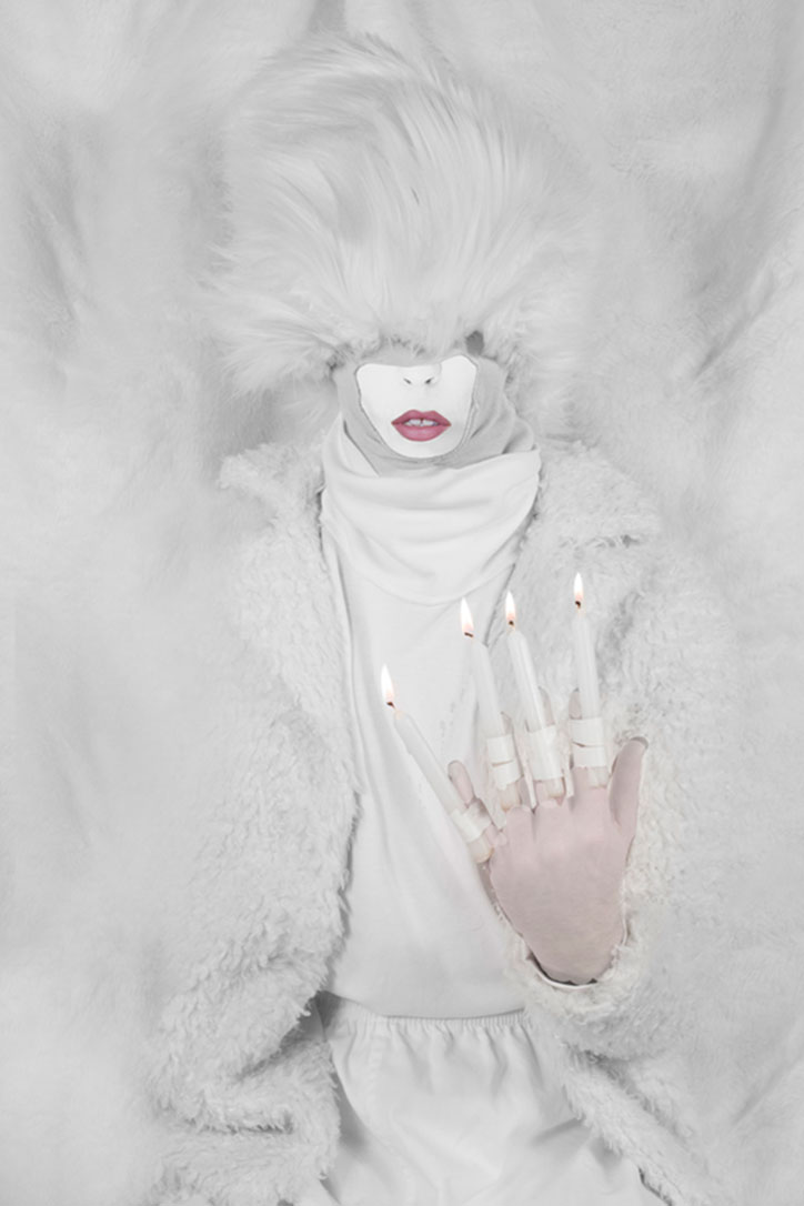 A person dressed entirely in white, including a fluffy coat and a large faux-fur headpiece that obscures their eyes. They hold up one hand with lit candles attached to four of their fingers.