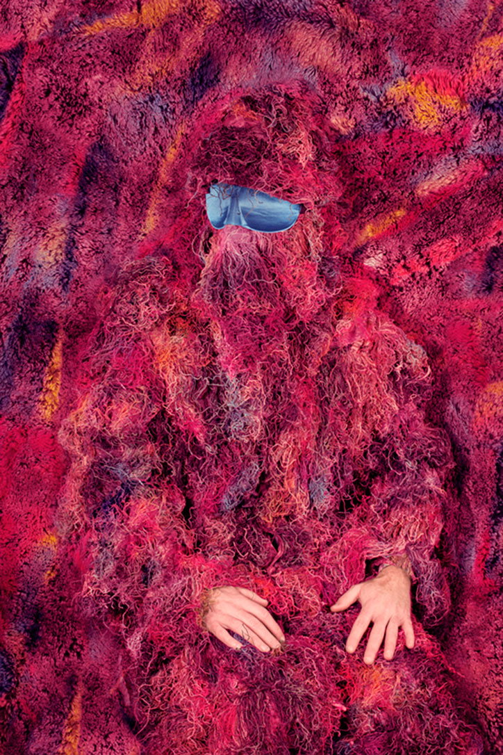 A person is completely enveloped in a voluminous, shaggy garment of deep pink and purple wool. A small piece of blue fabric covers their eyes, and their hands rest at their sides.
